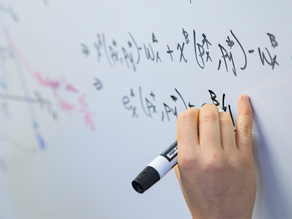 A hand holds a black dry erase marker while writing a series of formulas.